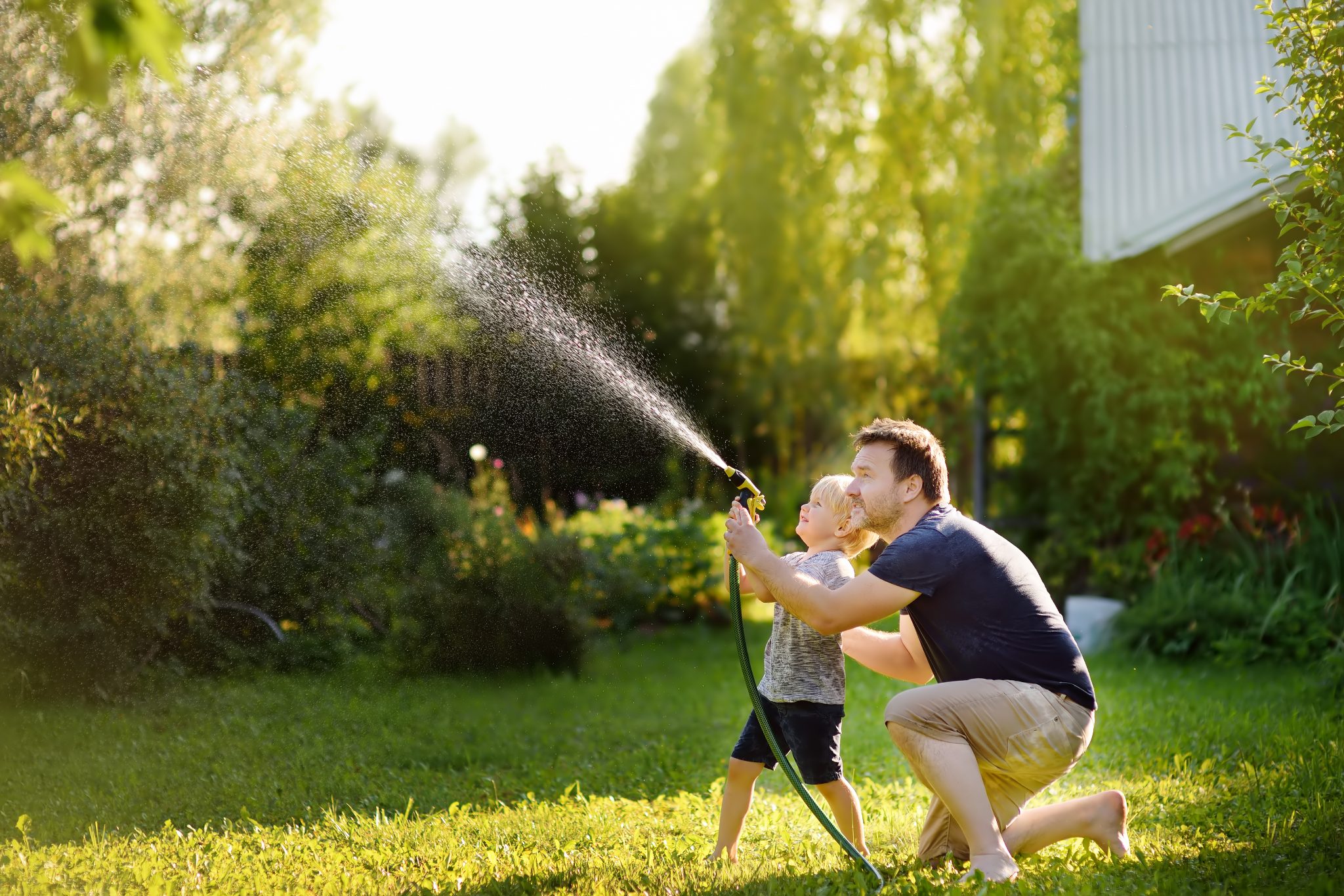 有趣的小男孩和他的父亲在阳光明媚的后院玩花园软管